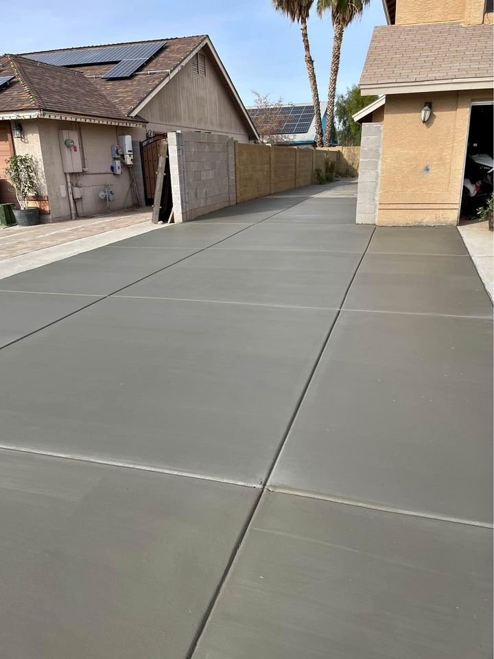 A freshly poured concrete driveway in a residential area, expertly crafted by a licensed and insured concrete contractor. The smooth, clean surface extends from the street up to a house with beige siding and a garage slightly visible on the right. The neighboring house has solar panels on its roof under the clear Phoenix sky.