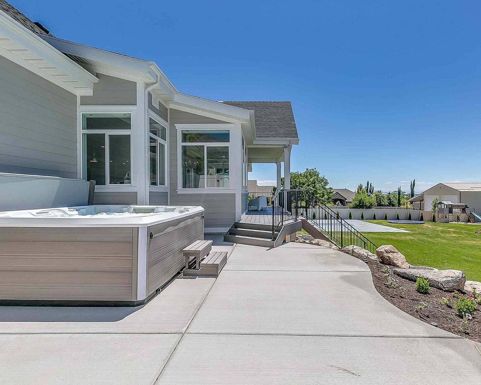 A spacious backyard in Phoenix AZ features a light grey house with a large windowed extension, a modern hot tub on a concrete patio by expert concrete contractors, and well-maintained landscaping. The scene includes a green lawn, stone accents, and a clear blue sky on a sunny day.