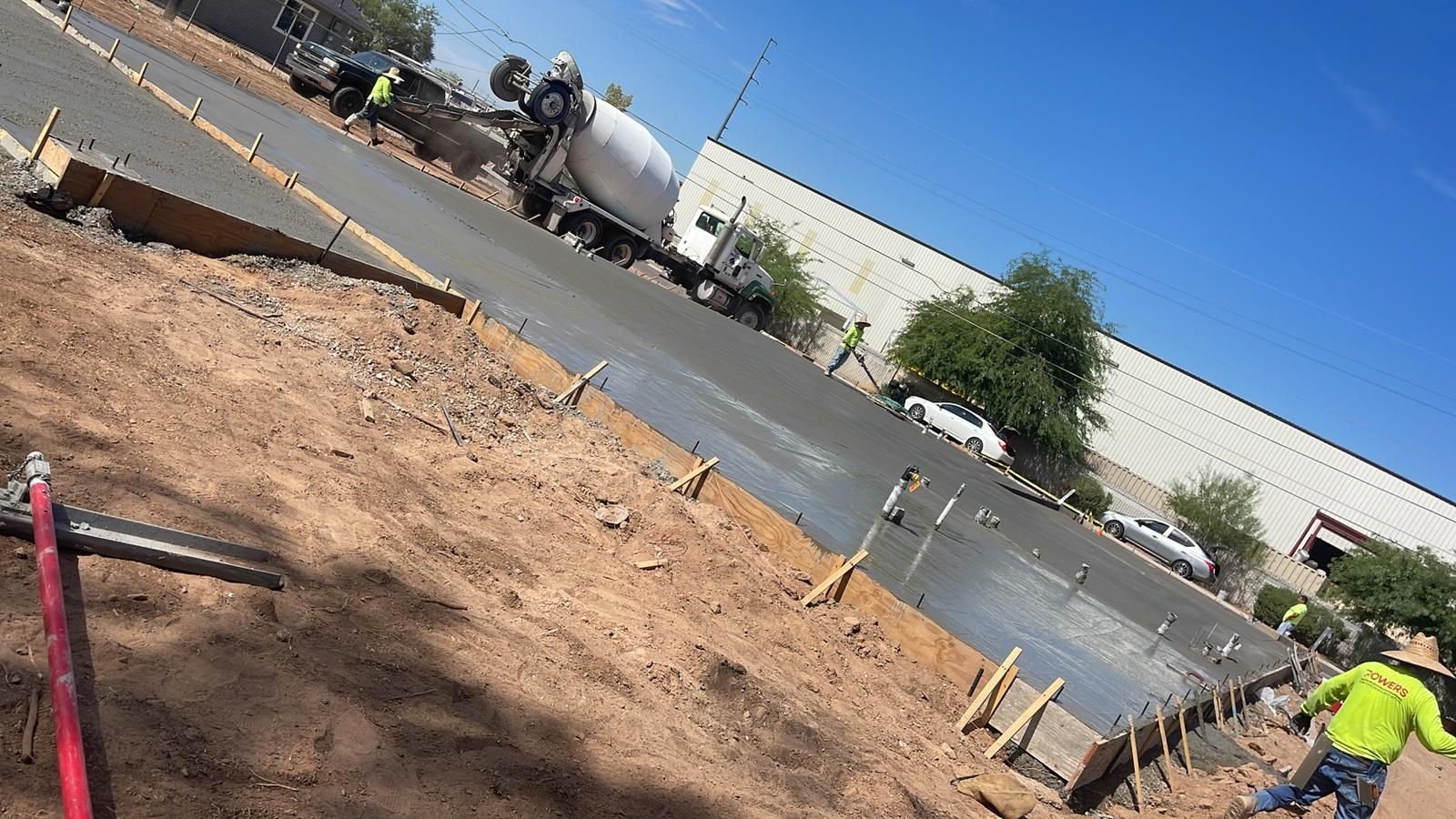 Construction site with workers pouring concrete from a cement truck into a large area framed with wooden planks. Several workers are visible managing the process, and cars are parked beside a large white building in the background under a clear blue sky. Concrete driveway installation is underway.