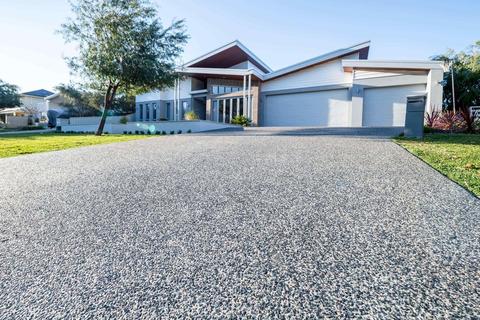 Modern house with large, sloped driveway leading to a three-car garage. The house features a unique angular roof design, full glass entry doors, and landscaped front yard with trees and shrubs. Expertly crafted by AZ Desert Concrete, the property basks in sunlight under a clear sky.