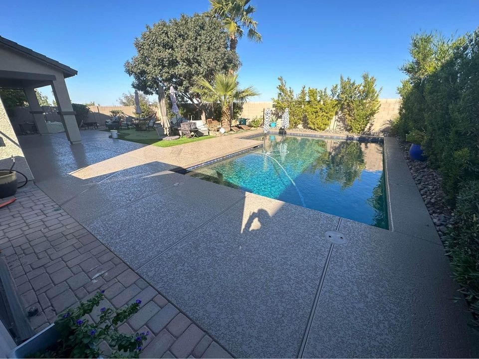 A backyard with a swimming pool surrounded by a concrete deck and a paver patio, expertly crafted by a licensed concrete contractor in the Phoenix Metro Area. Trees and bushes line the back fence. On the left, there's a covered patio area with outdoor furniture. The sky is clear and blue.