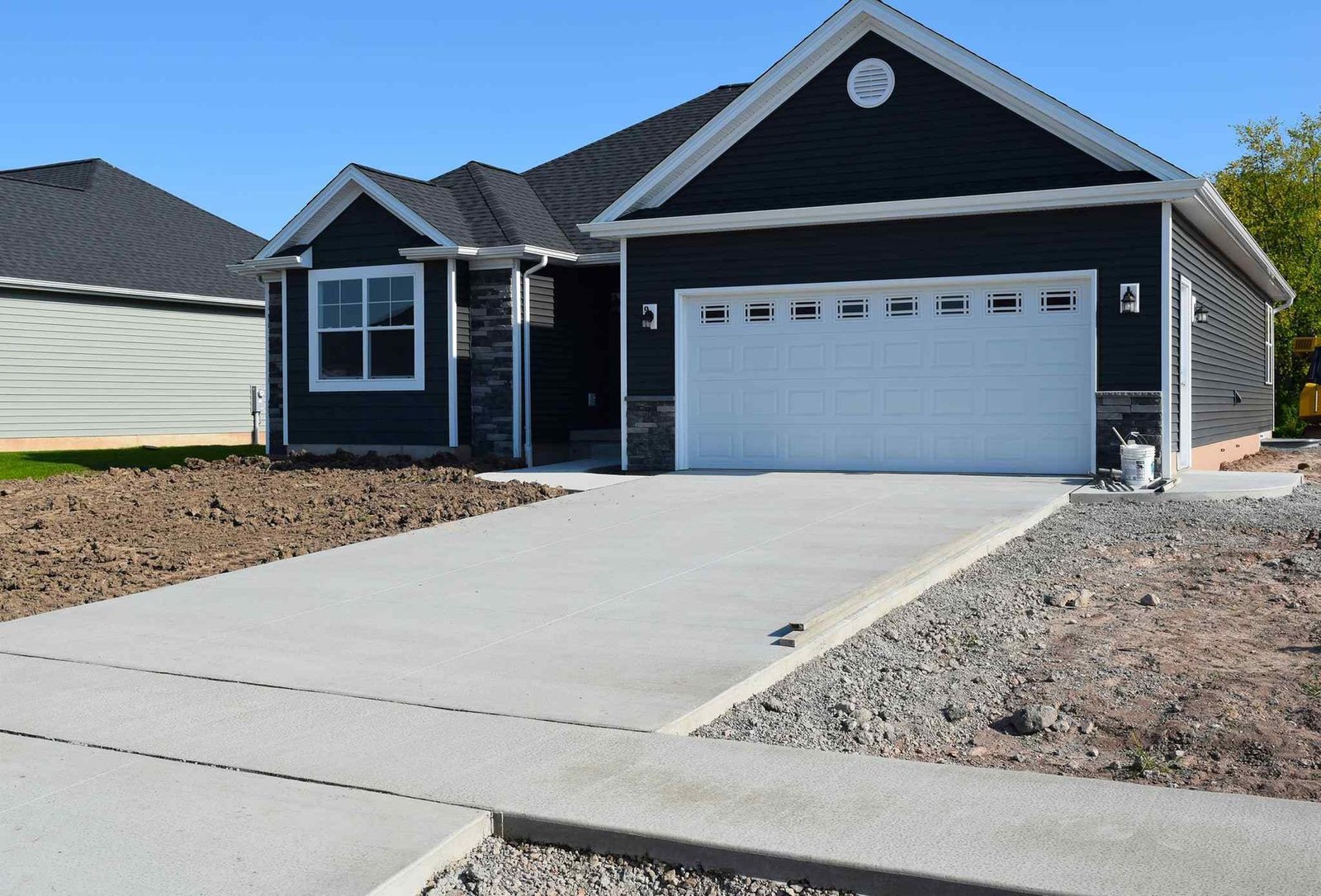 A single-story, dark gray house with white trim boasts a large two-car garage featuring a pristine white door. The front yard is mostly dirt with some construction materials left from the recent concrete driveway installation by certified installers in Phoenix, AZ. The sky is clear and blue.