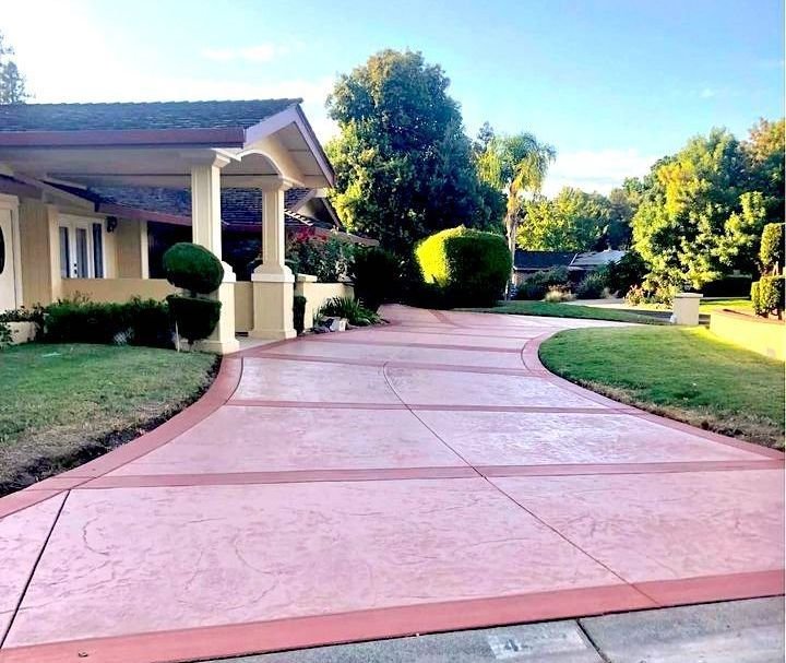 A suburban house with a well-maintained garden features a beige exterior and a covered porch. The wide, pinkish driveway, expertly crafted by a Phoenix concrete contractor, is lined with trimmed bushes and green grass on both sides, leading up to the main entrance. Trees are visible in the background.