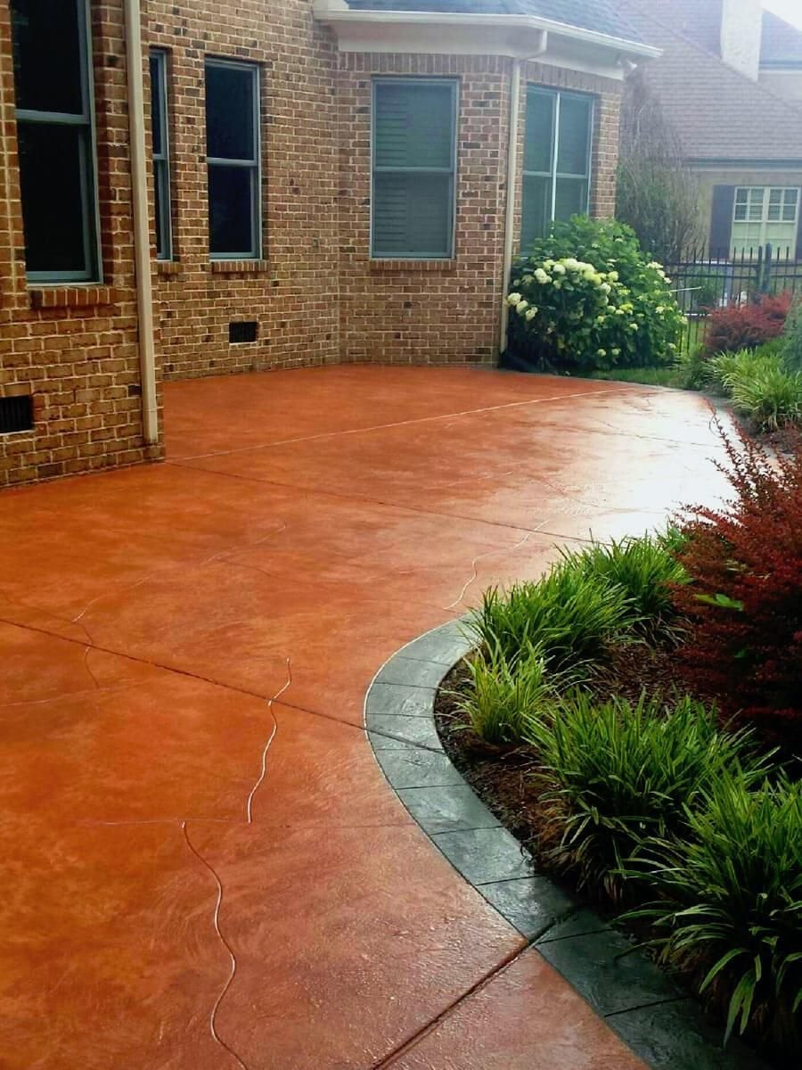 A curved brick pathway surrounds a well-maintained garden area with a variety of greenery and flowers. Adjacent to the garden is a large, red stamped concrete patio, expertly crafted by a licensed and insured Phoenix concrete contractor, extending from a brick house with windows and a backdoor.