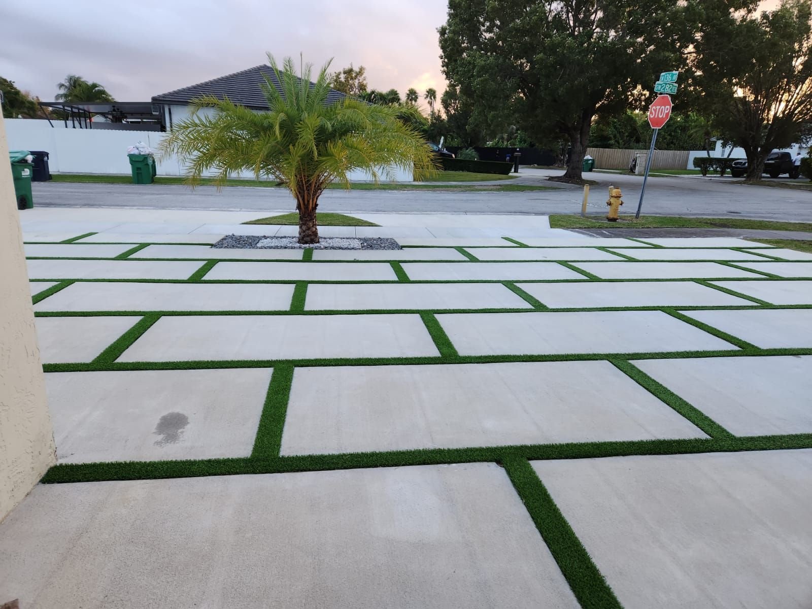 A modern driveway features a pattern of rectangular concrete slabs with strips of green grass in between. Installed by certified installers in Phoenix AZ, it boasts a small palm tree planted in a gravel bed at the center. A stop sign and fire hydrant are visible at the street corner, with trees and a white fence in the background.