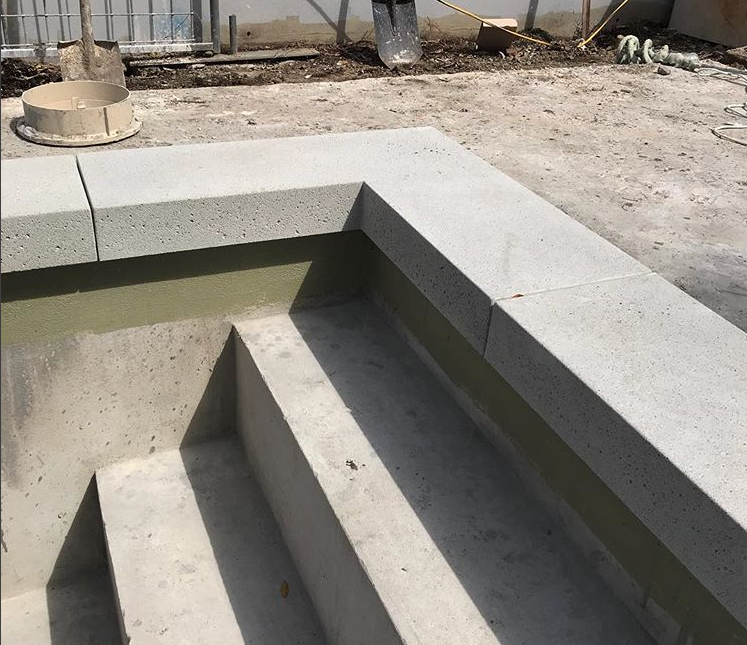 A concrete stairwell leading down into a partially built outdoor pool area in Phoenix. The unfinished pool's interior showcases exposed concrete surfaces, indicating an ongoing concrete pool deck installation. Construction materials and a shovel are visible in the background, hinting at the work of a skilled concrete installer.